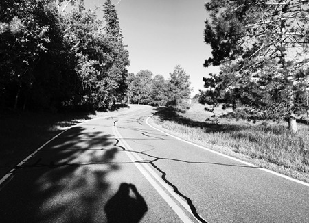 A cracked two-lane road curving through trees, with the photographer’s shadow in the foreground
