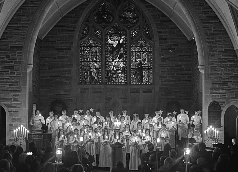 choir, lit by handheld candles, in front of a large stained glass window